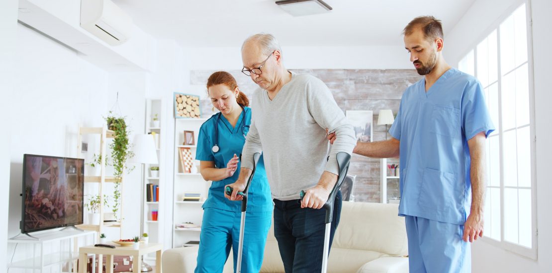 Team of nurses or social workers helping an old disabled man to walk with his crutches out of the nursing home room