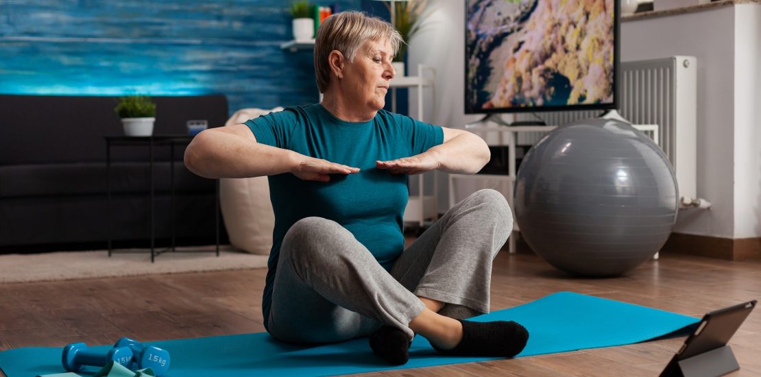Retired senior woman eatching fitness tutorial on laptop sitting on yoga mat streching arm