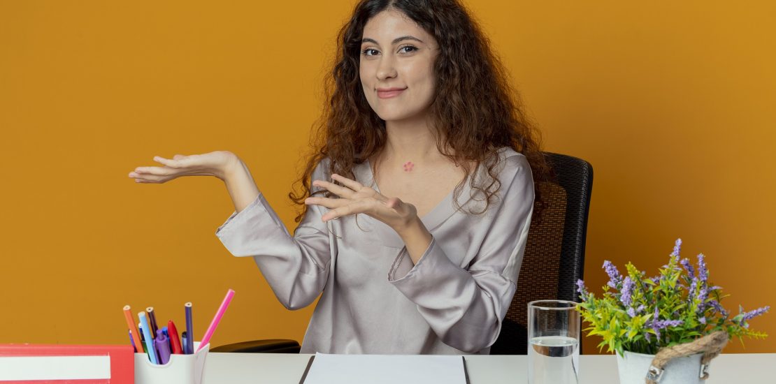 pleased young pretty female office worker sitting at desk with office tools points with hands at side isolated on orange background